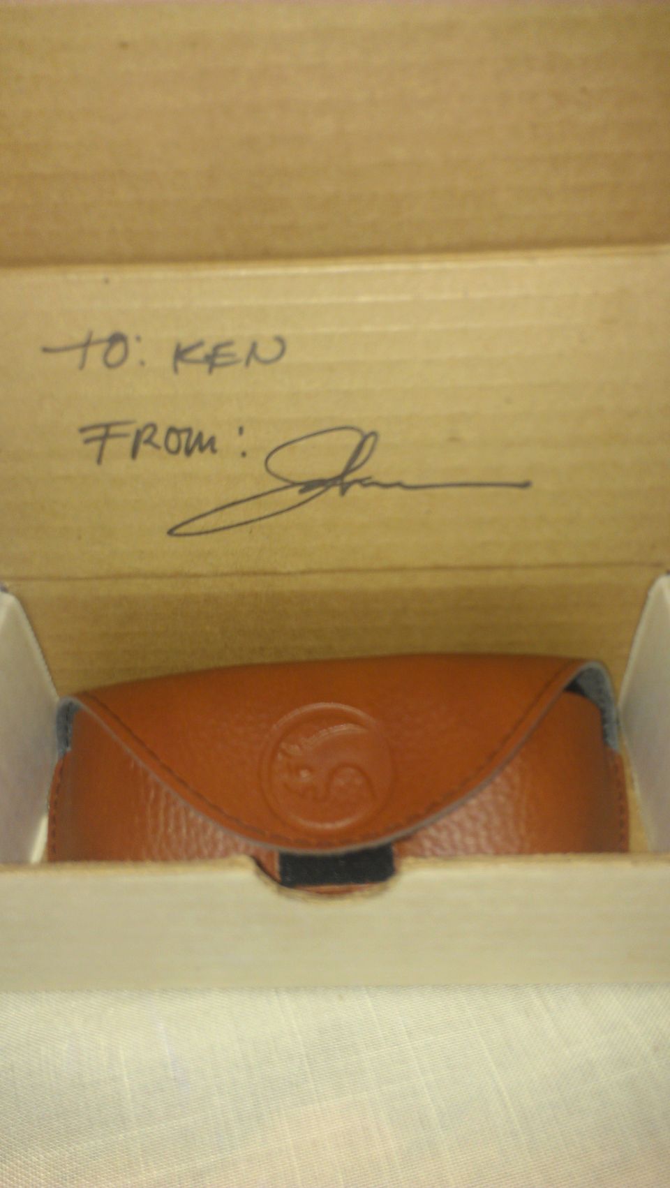 Brown leather glasses case with embossed logo, inside a cardboard box with handwritten 'TO: KEN FROM: John'
