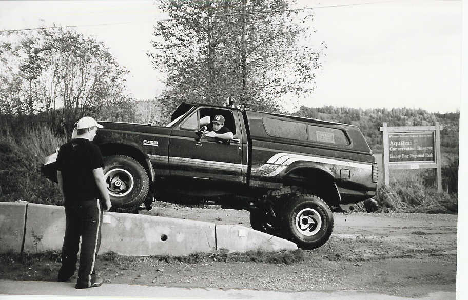 Dark Ford F-150 pickup truck with a camper shell, angled on a concrete barrier. A person is in the driver's seat