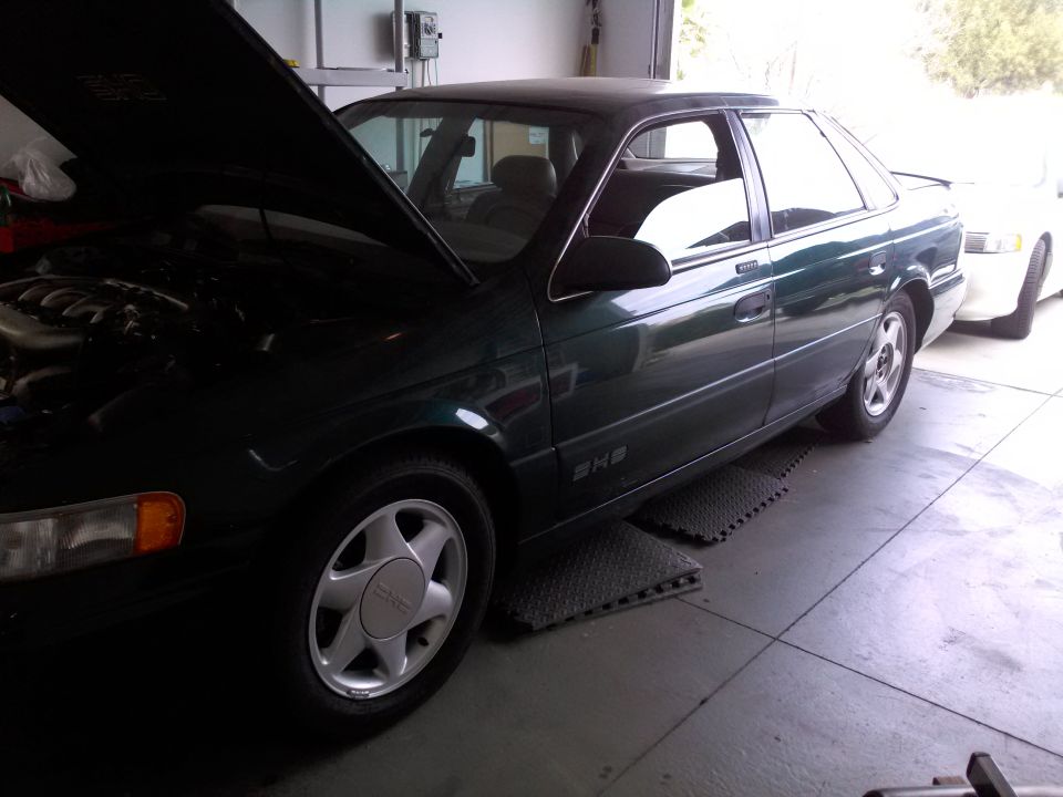 Green 1993 Ford Taurus sedan with hood open, parked next to a white car in a garage