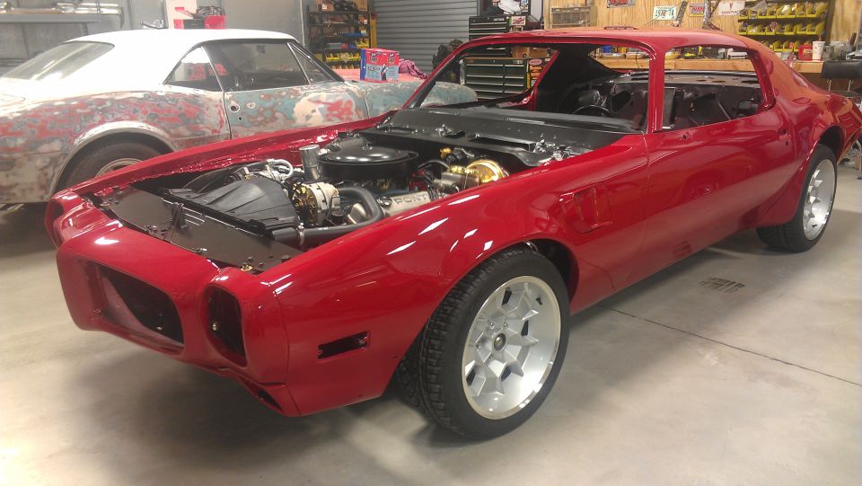 Red Pontiac Firebird with hood open, showing engine bay. Another car in primer is behind it