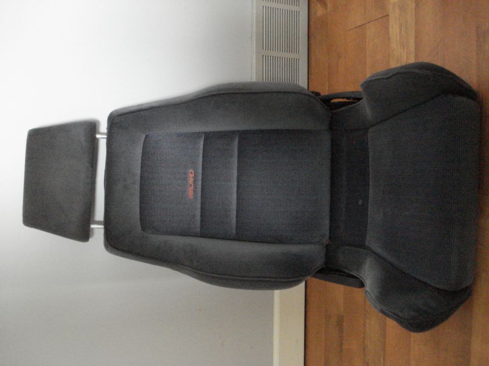 Dark gray fabric car seat with orange RECARO logo, detached headrest, on wooden floor