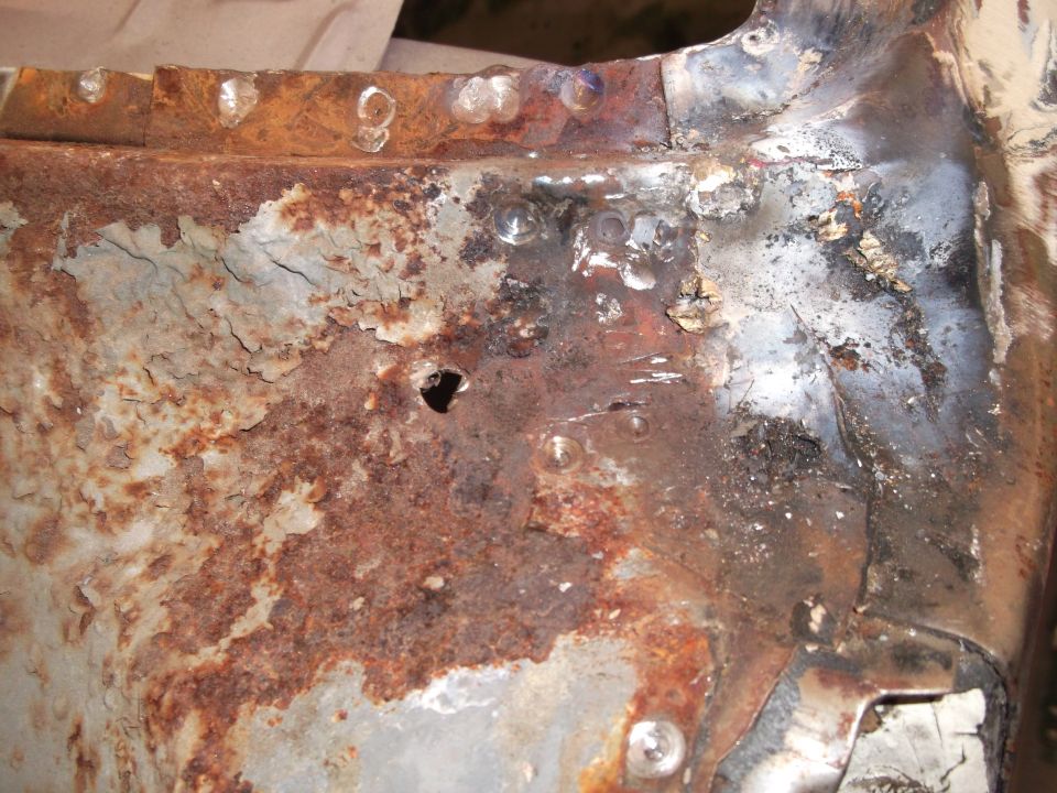 Heavily rusted metal panel with a hole, showing welds and peeling paint