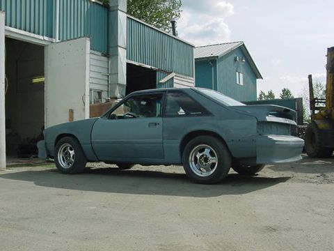 1990 Ford F-150 - Primer-finished 1990 Ford Mustang coupe sits outside a workshop with a forklift in the background.