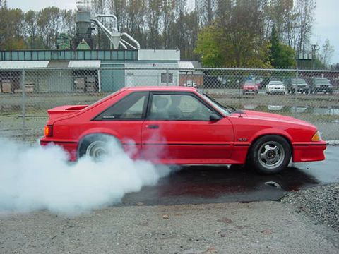 1990 Ford F-150 - A red coupe is seen from the side performing a burnout, with smoke billowing from its rear tires.