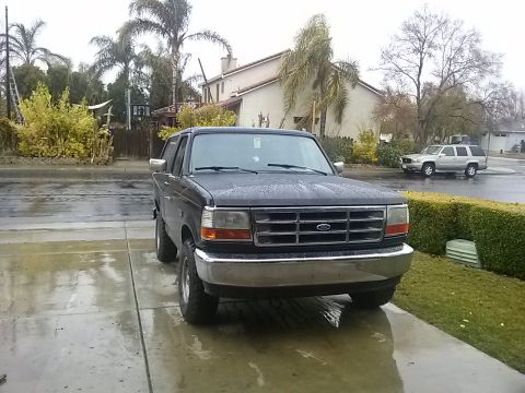 1996 Ford Bronco - Front view of a 1996 Ford Bronco parked on a wet driveway with a house and trees in the background.