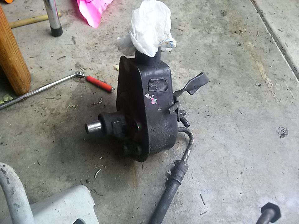 power steering pump sitting on concrete floor, with a rag stuffed in top opening. Tools and a jack stand are nearby