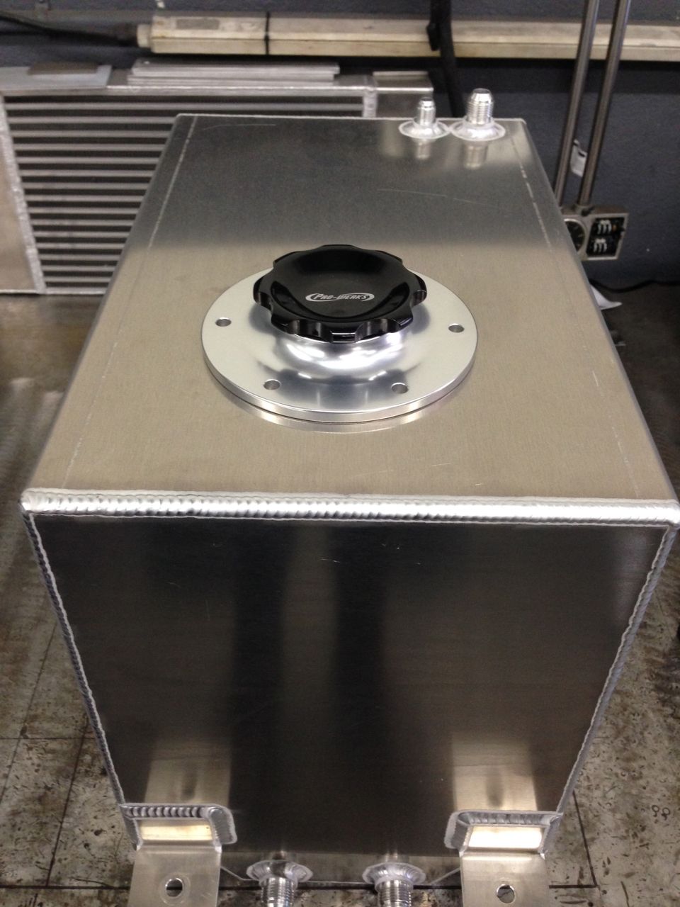 Aluminum fuel tank with black cap and two fittings on top, and two fittings on the bottom. Welded seams visible