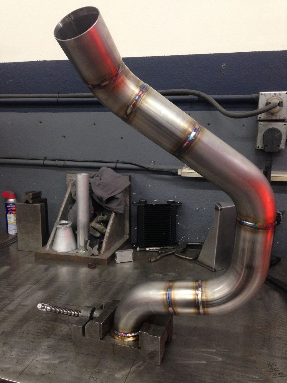 Custom bent stainless steel exhaust pipe with visible welds, clamped in a vise