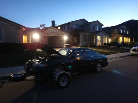1993 Ford Taurus - Dark sedan with hood open, flat tire, and a trailer attached, parked on a residential street at dusk.