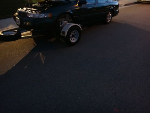 1993 Ford Taurus - 1993 Ford Taurus with its hood up and a trailer hitched to the front, spare tire nearby.