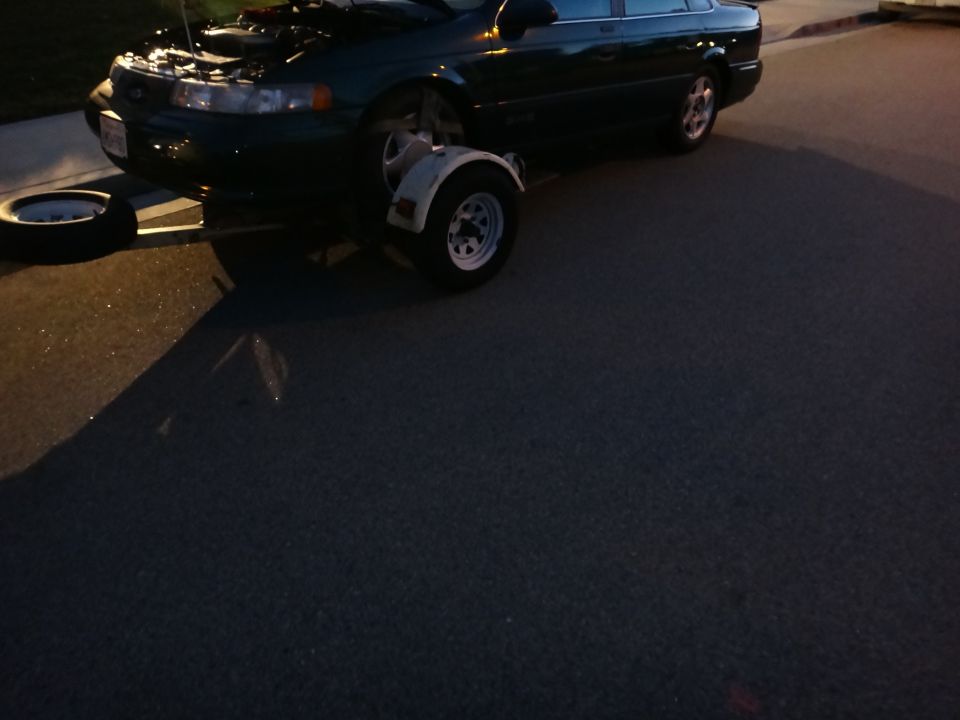 1993 Ford Taurus with hood open, a small trailer attached to the front bumper area, and a spare tire on the ground