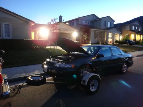 1993 Ford Taurus - A dark sedan is parked on a trailer with its hood open in a suburban neighborhood at dusk.