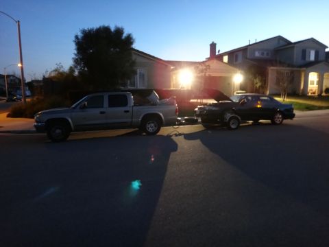 1993 Ford Taurus - A dark sedan with its hood open is parked on a street next to a pickup truck, with houses in the background.
