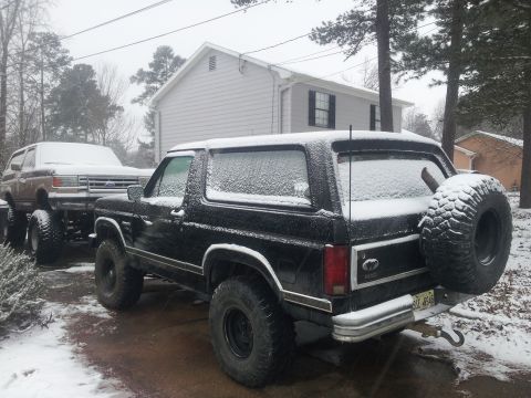1986 Ford Bronco - Two Ford Broncos are parked outside in the snow. The black Bronco has a spare tire mounted on the rear.
