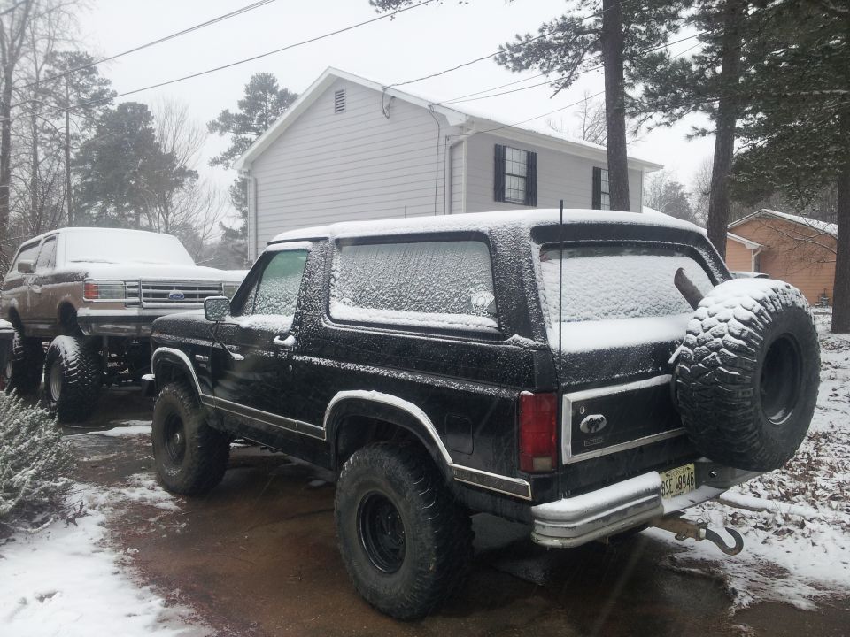 Two Ford Broncos parked in snow, one black with spare tire on back, another brown on trailer