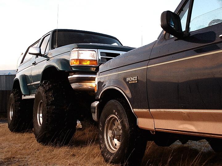 Two lifted Ford Broncos, teal and dark blue, on big mud tires in a grassy field