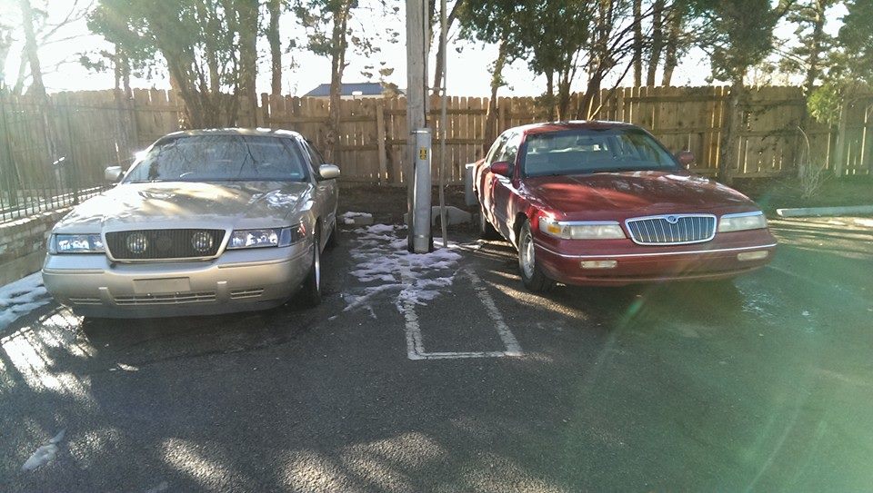 Two Mercury Grand Marquis sedans parked side-by-side. The one on the left is tan, and the one on the right is red. Both are facing forward