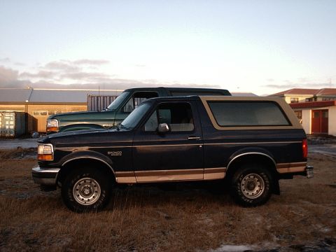 1996 Ford Bronco - Two SUVs, a dark blue Ford Bronco and a green truck, are parked side-by-side outdoors.