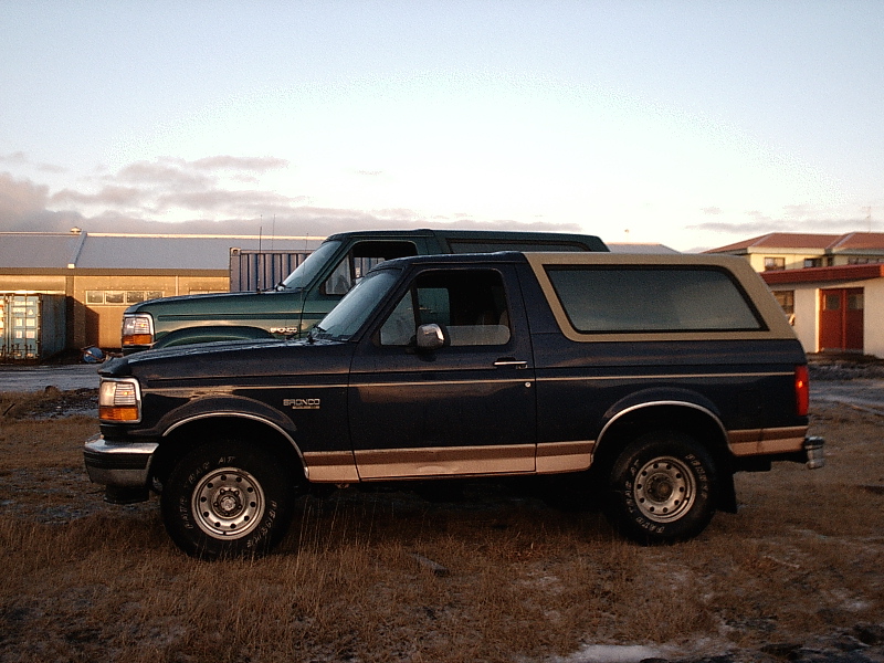 Dark blue Ford Bronco SUV with tan top, side view on gravel lot at dusk