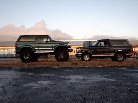 1996 Ford Bronco - Two Ford Broncos, a lifted green model and a dark blue one, are parked side-by-side under a cloudy sky.