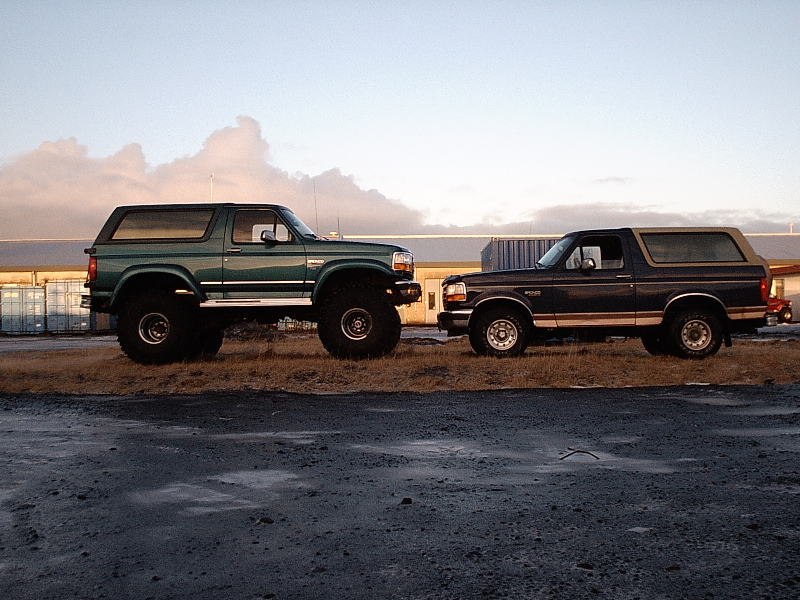 Green lifted Ford Bronco beside a dark blue stock Bronco, both in side view in a gravel lot