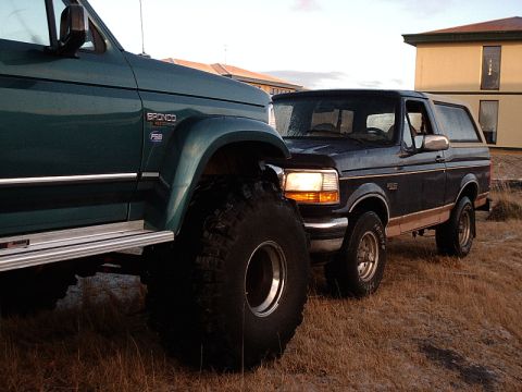 1996 Ford Bronco - Two Ford Broncos, one stock and one heavily modified with oversized tires, are parked side-by-side.