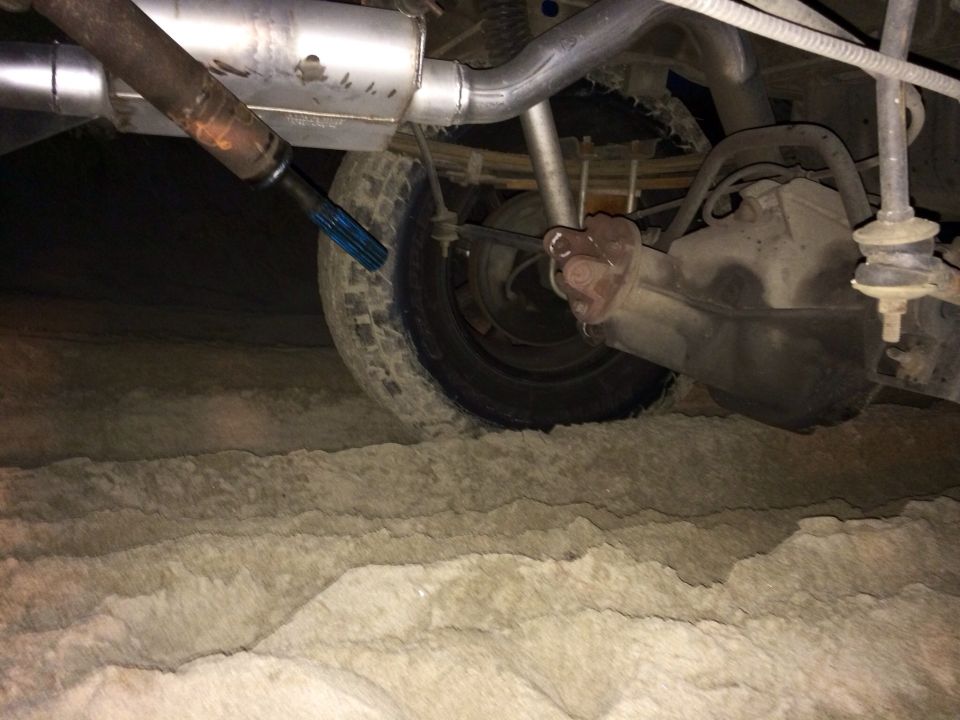Rear axle, driveshaft, and exhaust of a vehicle in sand. The driveshaft has a blue protective cap