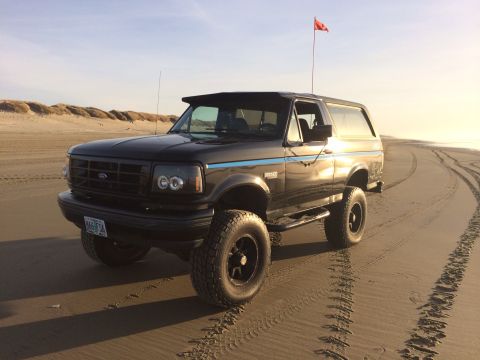 1992 Ford Bronco - A black Ford Bronco is parked on a sandy beach near sand dunes under a clear sky.