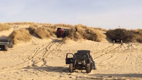 1992 Ford Bronco - Multiple off-road vehicles navigate sandy terrain with one vehicle overturned on a dune.