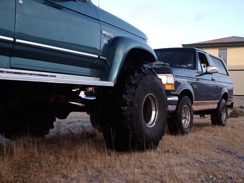 1996 Ford Bronco - Two Ford Broncos are parked side-by-side on a dry, grassy field with a building in the background.