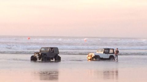 1992 Ford Bronco - Two Jeep Wranglers are seen driving on a beach as the sun sets over the ocean.