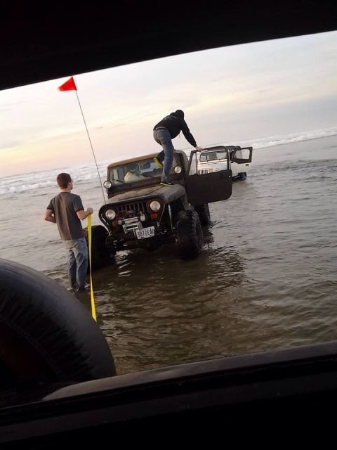 1992 Ford Bronco - Two Jeeps are stuck in the water on a beach, with people interacting with them.