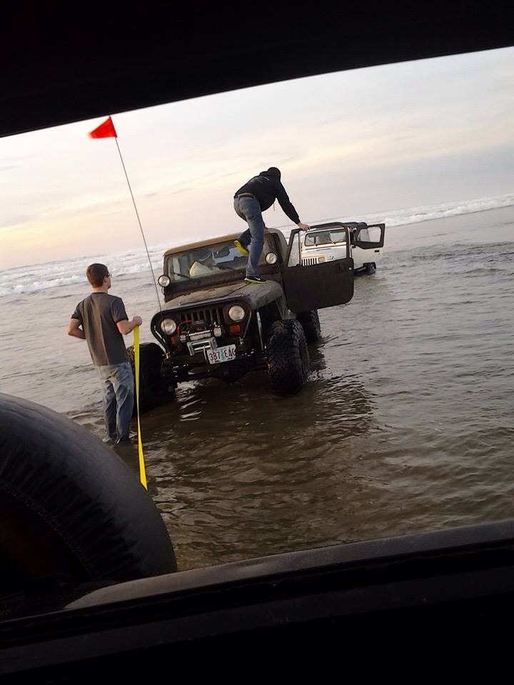 Two Jeeps partially submerged in water on a beach, one with a person climbing on it and another with the door open
