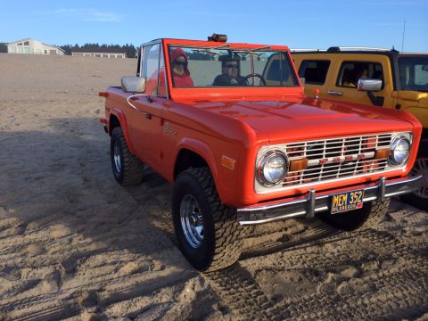 1992 Ford Bronco - This orange convertible SUV is parked on a sandy beach, with another yellow vehicle visible behind it.