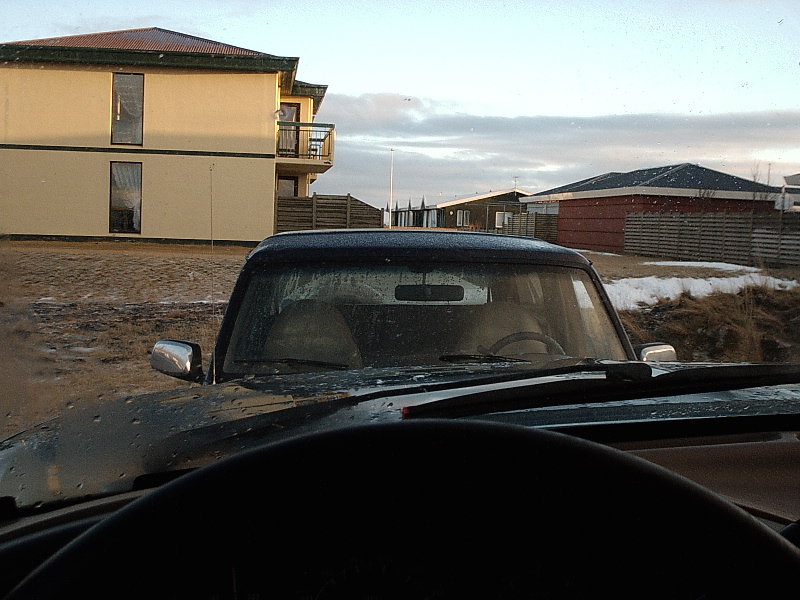 Dark SUV seen through windshield in a snowy muddy lot near buildings