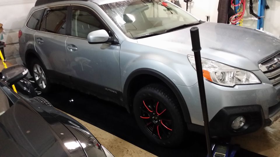 Silver 2014 Subaru Outback parked indoors with black wheels and red accents. Another car is partially visible in the foreground