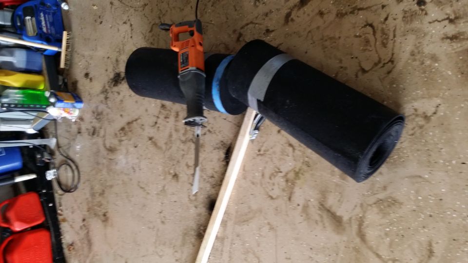 Reciprocating saw cutting a rolled black mat secured with tape and a wooden brace
