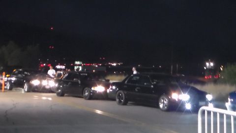 2003 Ford Crown Victoria - nighttime scene of several dark cars with headlights on lined up at a track staging area, with people standing nearby and lights in the dis…