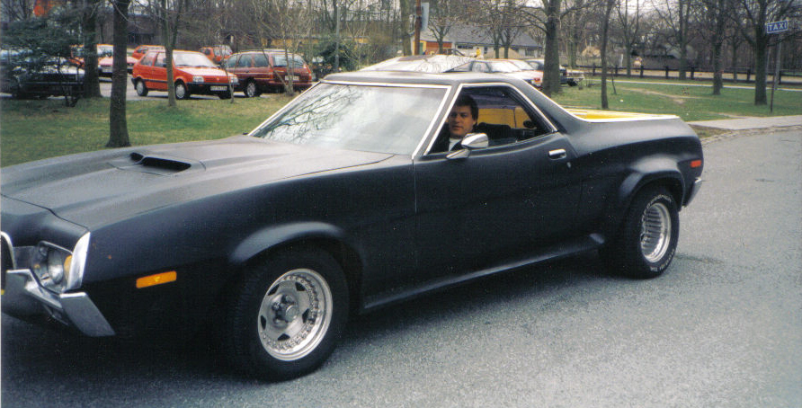 Black custom pickup truck with a scoop on the hood, driver inside. Other cars parked in background