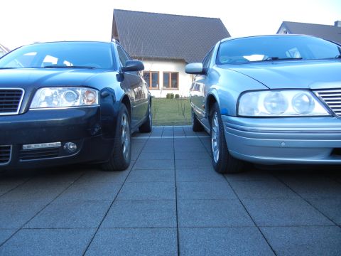 1999 Ford Fairlane - Two cars are parked side-by-side on a paved driveway in front of a house.