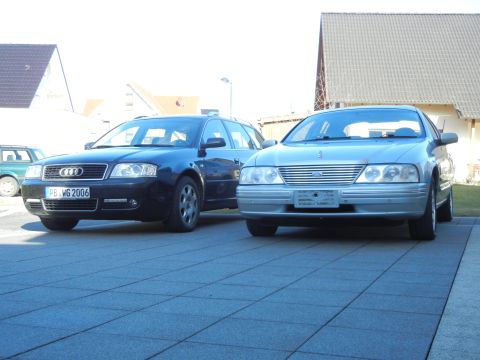 1999 Ford Fairlane - Two cars are parked on a tiled surface in front of houses.