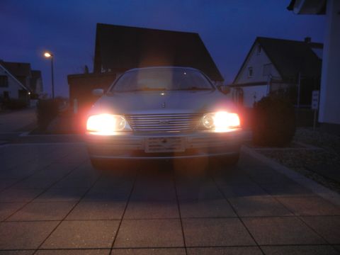 1999 Ford Fairlane - A silver sedan is parked in a driveway at dusk, its headlights illuminating the scene.