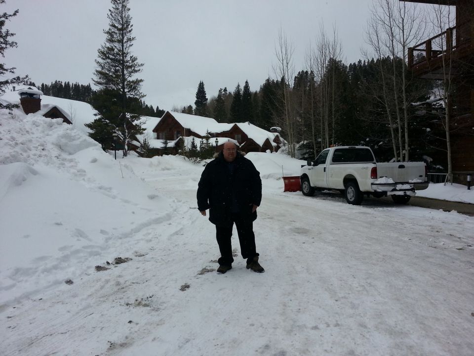 White pickup truck with a red snow plow attachment parked in a snowy area with buildings and trees