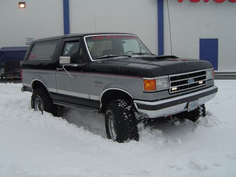 1989 Ford Bronco - A two-tone Ford Bronco navigates through deep snow, its front grille and headlights clearly visible.