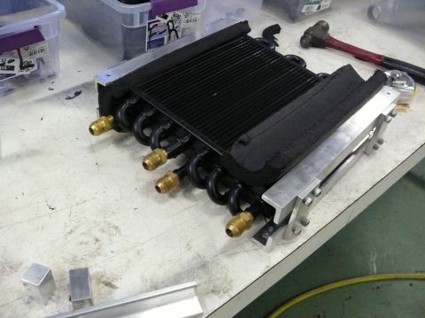 1976 Chevrolet Corvette - A transmission cooler with black fins and coiled lines sits on a cluttered workbench next to a hammer.