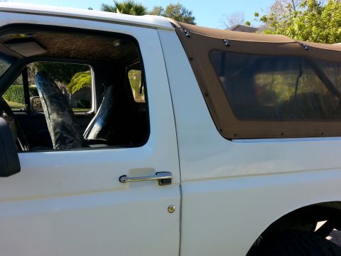 1992 Ford Bronco - The side profile of a white 1992 Ford Bronco reveals its interior and a brown soft top, with camouflage seat covers visible.