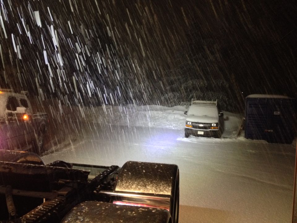 Truck cab and wheels in foreground, white van in background during snowfall