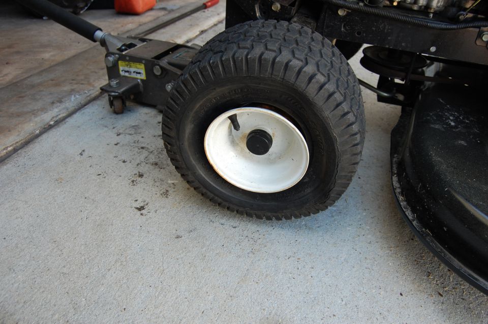 Tire on a white rim, lifted by a hydraulic jack, next to a black lawn mower deck