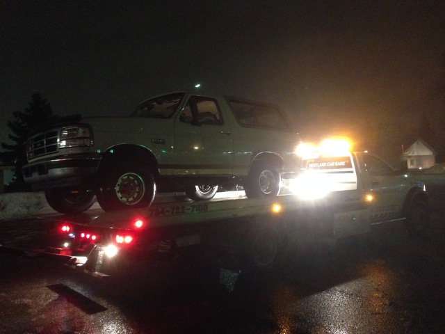 1995 Ford Bronco on a flatbed tow truck at night, with lights on the tow truck