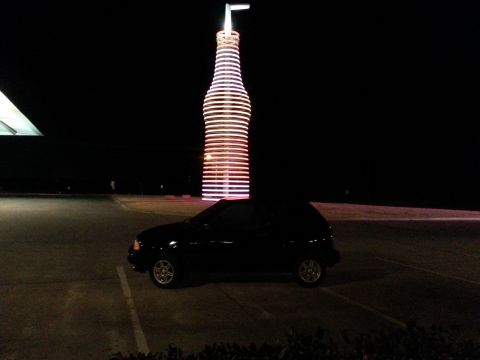 1991 Ford Festiva - A black hatchback is parked in a lot at night, with a giant illuminated bottle sign behind it.